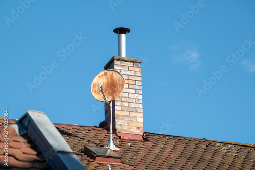 Untersicht verwitterte Satellitenschüssel und gemauerter Schornstein aus Backsteinen mit Metallrohr auf Ziegeldach  in der bürgerlichen Bergstadt in Bamberg vor blauem Himmel.	
