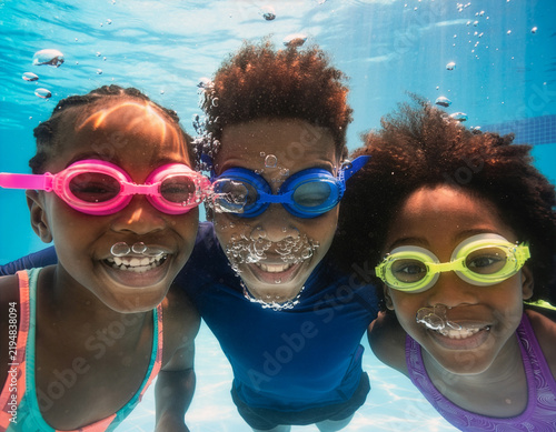 Group of cheerful children, girls and boy, are wearing swimming goggles and happily looking at the camera underwater in a blue swimming pool, with bubbles all around them, enjoying a relaxing spa.