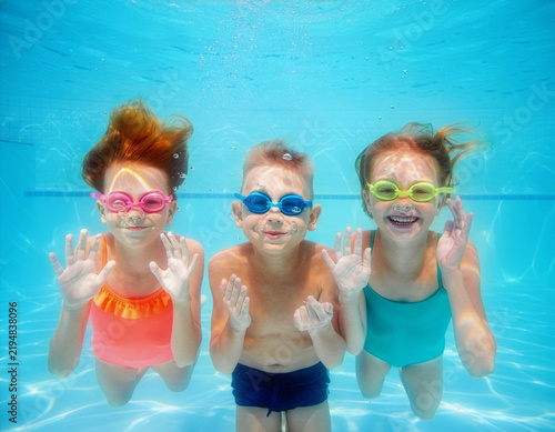 Group of cheerful children, girls and boy, are wearing swimming goggles and happily looking at the camera underwater in a blue swimming pool, with bubbles all around them, enjoying a relaxing spa.