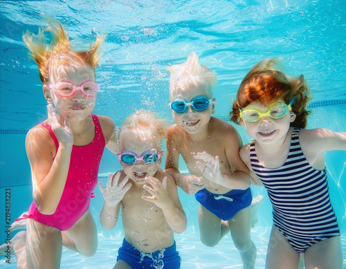 Group of cheerful children, girls and boy, are wearing swimming goggles and happily looking at the camera underwater in a blue swimming pool, with bubbles all around them, enjoying a relaxing spa.
