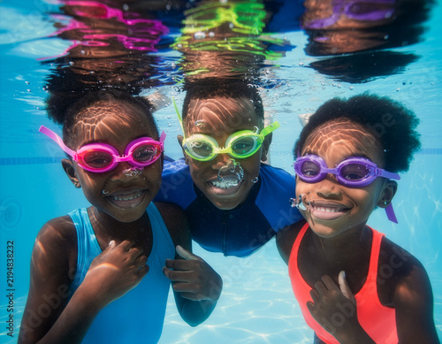 Group of cheerful children, girls and boy, are wearing swimming goggles and happily looking at the camera underwater in a blue swimming pool, with bubbles all around them, enjoying a relaxing spa.