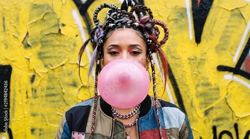 A stylish young woman with elaborate braids blows a large pink bubblegum bubble in front of a vibrant, graffiti-covered wall, embodying a bold and youthful spirit.
