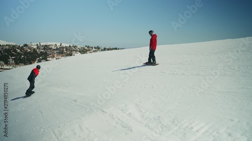 Two snowboarders stand on a sunny, snow-covered mountain slope with a distant resort town. This outdoor winter sport scene showcases active leisure and the joy of snowboarding.