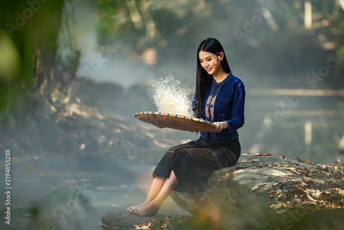 Smiling barefoot young woman in traditional Thai clothing sitting on a rock by a river winnowing rice (Fad Khao) using a bamboo tray (Kradong), Sakon Nakhon, Thailand
