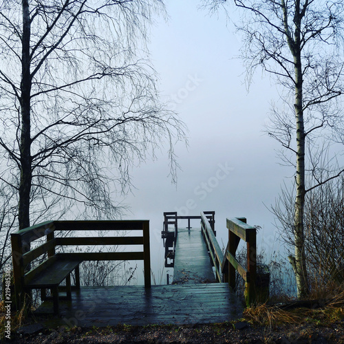 A wooden jetty with a bench overlooks a calm and misty lake surrounded by birch trees.