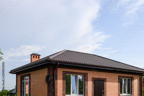 New single-story brick house under construction with finished tiled roof, chimney, gutters and installed windows, showcasing modern residential building and exterior construction