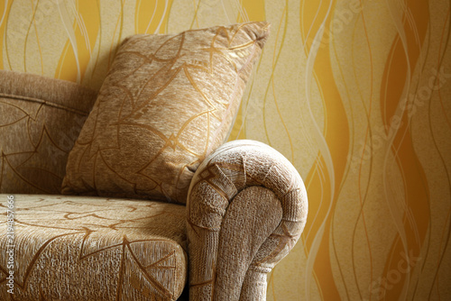 Close-up view of cozy upholstered sofa with decorative cushions and rounded armrest in warm brown and golden tones. Textured fabric, geometric patterns, soft lighting create comfortable room interior