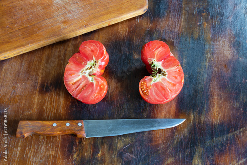 Two halves of unusually shaped, ugly red tomato lie on rustic wooden table after being cut open with kitchen knife. Cut surfaces reveal fleshy interior, seed chambers, and irregular natural structure