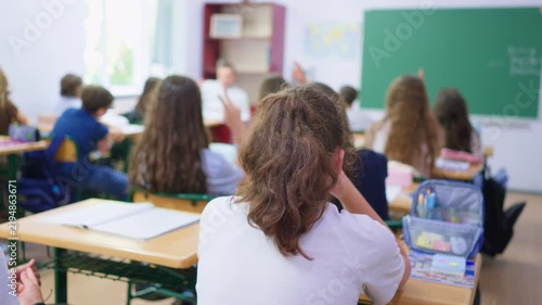 A group of young students sits at wooden desks in a bright classroom, while a classmate writes lesson notes on the large green chalkboard at the front of the room