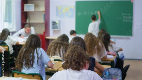 A group of young students sits at wooden desks in a bright classroom, while a classmate writes lesson notes on the large green chalkboard at the front of the room