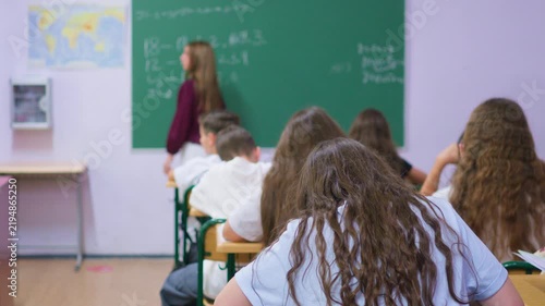 A group of young students sits at wooden desks in a bright classroom, while a classmate writes lesson notes on the large green chalkboard at the front of the room