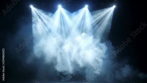 Four bright spotlights illuminating a smoky stage with blueish-white beams of light against a dark background