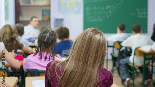 A group of young students sits at wooden desks in a bright classroom, while a classmate writes lesson notes on the large green chalkboard at the front of the room
