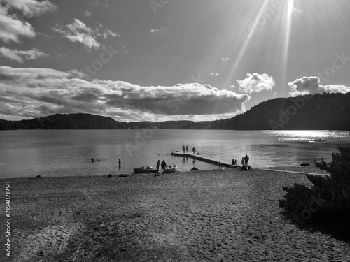 Black and white view of people gathered on a wooden pier and sandy beach on a sunny day by a lake.