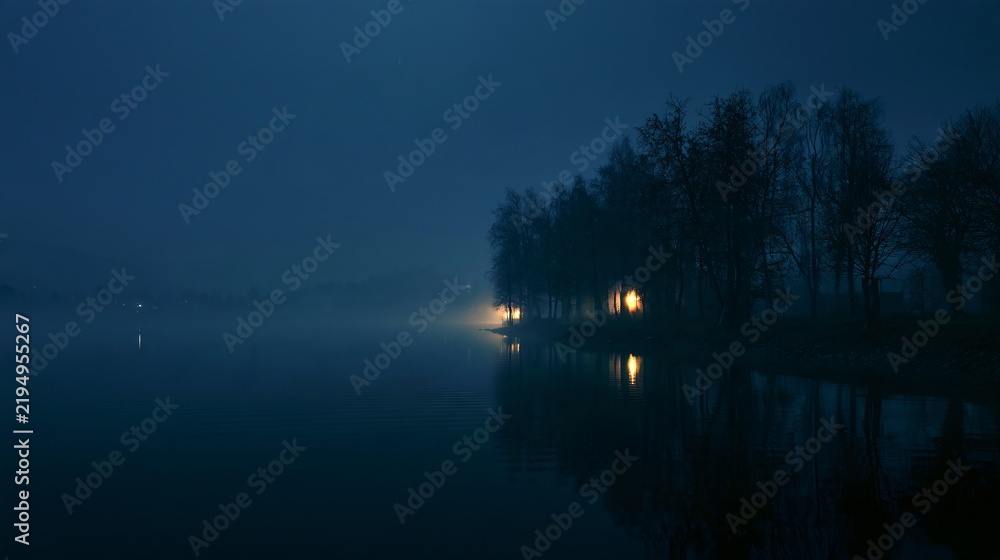 Obraz premium A Halloween background showing a foggy lake at night with the faint silhouette of leafless trees along the shoreline and a few distant warm lights reflected in the water, deep navy atmosphere