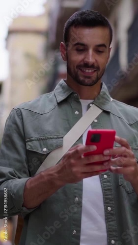 Cheerful young man walking towards the camera while using smartphone to send online messages and check social media app. Vertical video.