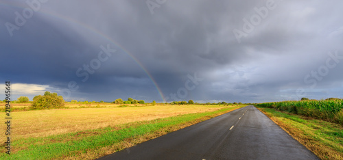 Bild auf Leinwand Storm clouds break as rainbow appears over rural road