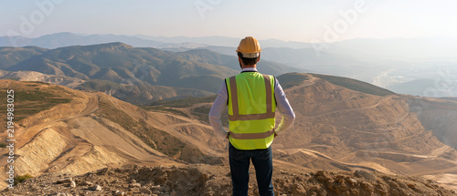 Miner Surveying Vast Landscape: A lone miner, clad in safety gear including a hard hat and reflective vest, surveys a vast, open-pit mining landscape under a clear sky.