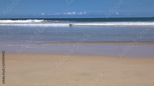 A dramatic seascape showing powerful ocean waves breaking and splashing near a small rocky island. The deep blue sea stretches to the horizon under a clear blue sky, creating a vivid 