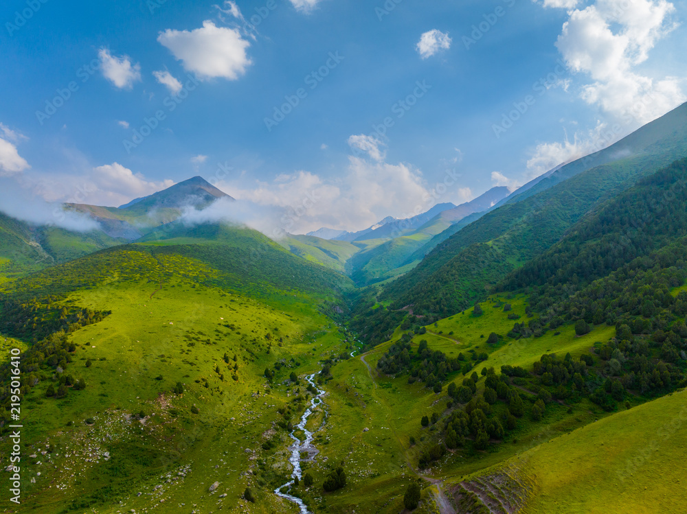 Obraz premium Aerial perspective reveals a breathtaking mountain valley bathed in daylight. A clear, shallow river snakes through the vibrant green landscape, flanked by rolling hills and steep mountain slopes.