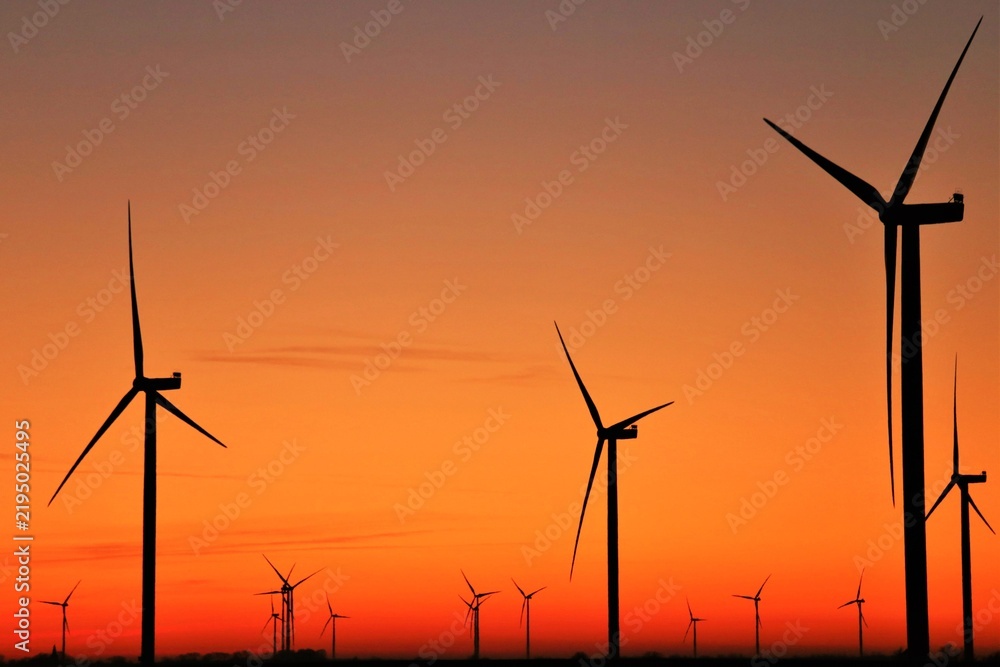 Obraz premium Vue panoramique d'un champ d'éoliennes dessinant des silhouettes verticales au coucher du soleil, sous un ciel orange mauve. Symbole d'une énergie renouvelable grâce à la force du vent.