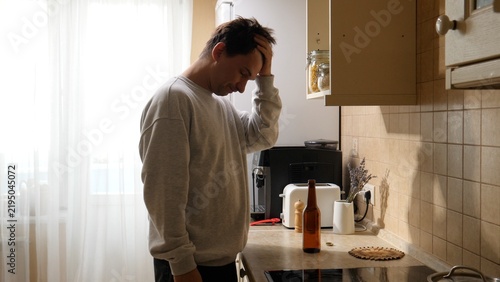 Canvastavla Man in kitchen clutching his head over an empty beer bottle, tired and distresse