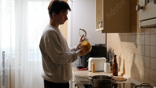 Canvastavla Young man in a modern home kitchen holding a glass jar of dry pasta while prepar
