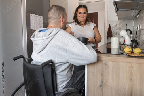 Female personal assistant helps a person in a wheelchair with meal preparation in a kitchen. They work together for independent living.