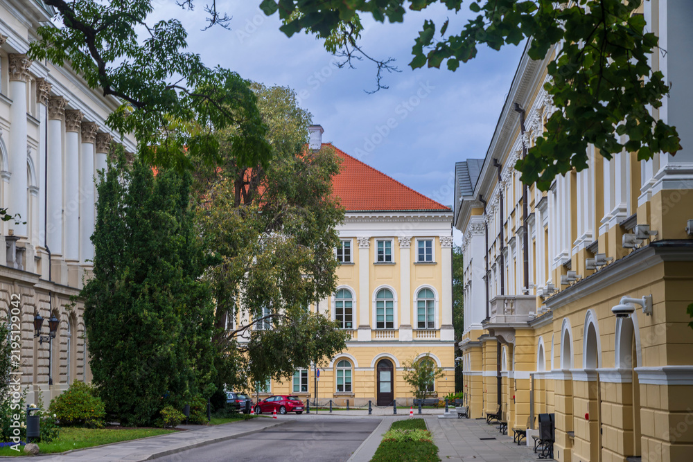 Obraz premium Warsaw, Poland Quiet inner street and courtyard surrounded by historic university campus buildings with yellow and white neoclassical facades. Green trees framing the view on an overcast day.