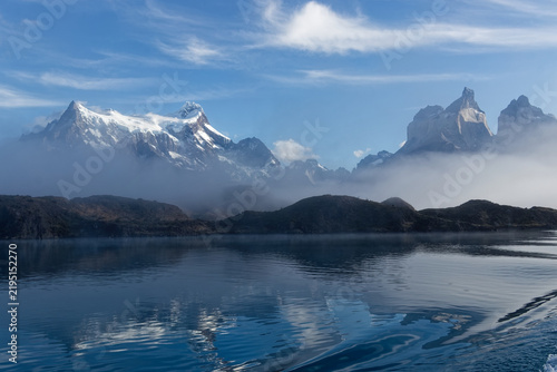 Snow covered peaks of Torres del Paine rising above morning fog and turquoise glacial waters of Lake Pehoe in Chilean Patagonia.