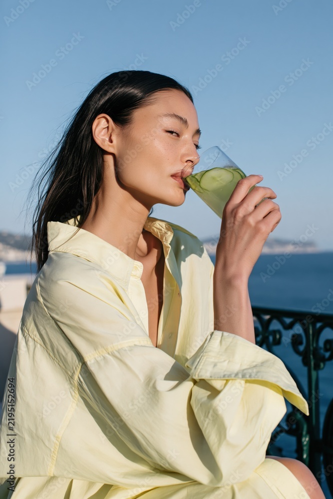 Fototapeta premium Profile of a woman sipping green juice with ocean view in an oversized yellow shirt for summer wellness lifestyle and travel banner