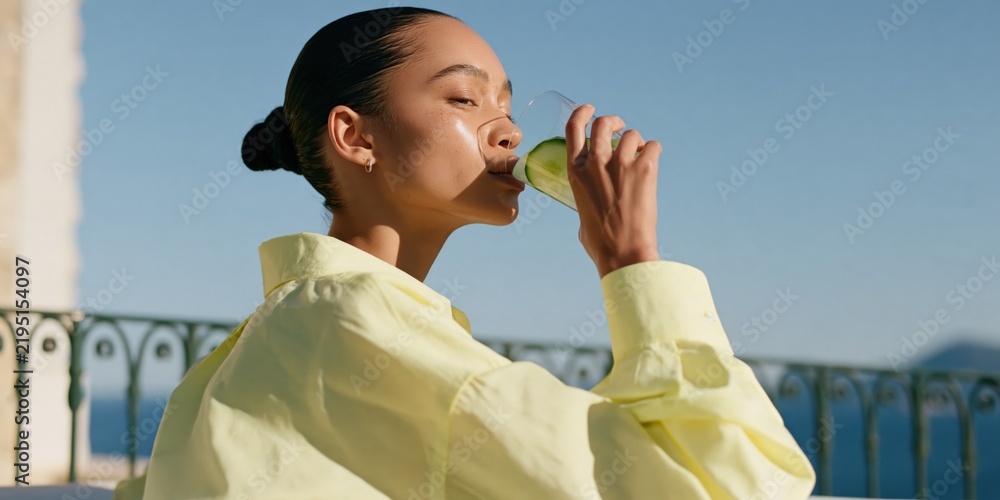 Fototapeta premium Young woman drinking green juice on balcony in sunlight wearing yellow shirt for wellness lifestyle banner
