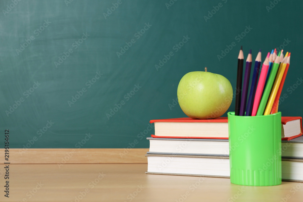 Obraz premium Stack of books, stationery and apple on wooden table against chalkboard. Space for text
