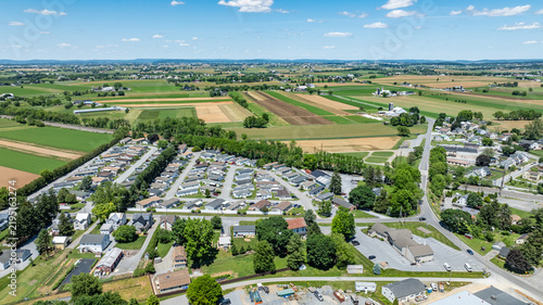 Aerial view shows fields and Mobile, Manufactured homes in a rural area. There are green and brown fields alongside a small neighborhood. The scene captures the layout of roads and houses.