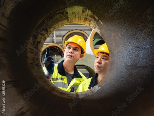 Asian Inspect Steel Coil Framed Through Industrial Pipe : factory engineer wearing yellow hardhat high-visibility jacket inspect warehouse inventory seen through round open industrial steel cylinder.