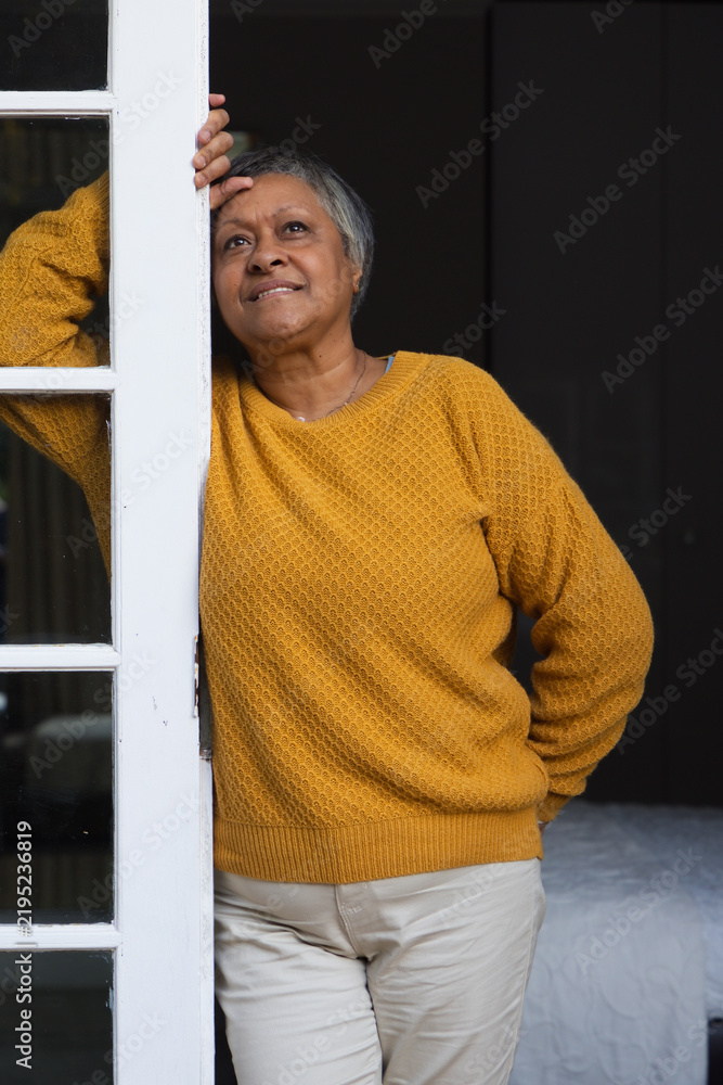 Obraz premium Senior African American woman leaning on white door at bedroom doorway in mustard sweater
