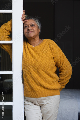 Senior African American woman leaning on white door at bedroom doorway in mustard sweater