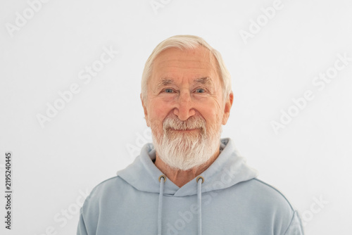 Obraz na płótnie Senior man in light hoodie stands against plain white studio wall, senior generation shown without cliche, emotion easy to recognise
