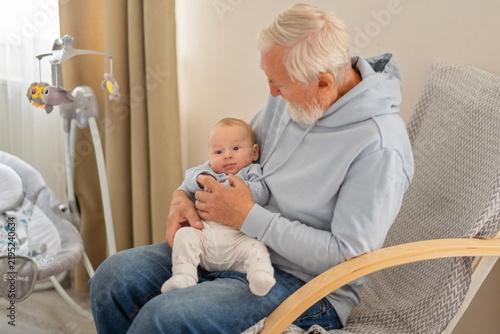 Obraz na plátně Elderly man sits with baby in nursery beside electric swing, generation gap turning into warm contact, mood for family campaign