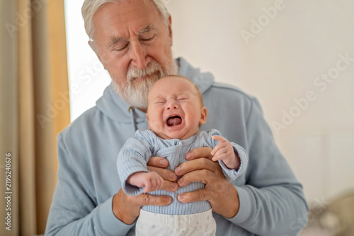 Obraz na płótnie Elderly man closes eyes while holding loudly crying grandson, soothing crying child with calm hands, emotion impossible to stage