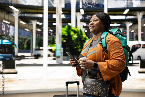 Canvastavla Smiling woman holds a mobile phone and coffee cup in a bus station, checking route details in the shuttle terminal while enjoying a morning beverage before departure and weekday travel