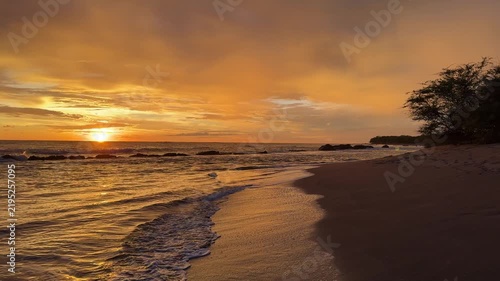 The sun sets on the horizon at a beach with calm waves moving ashore and a few small rocks in the water.