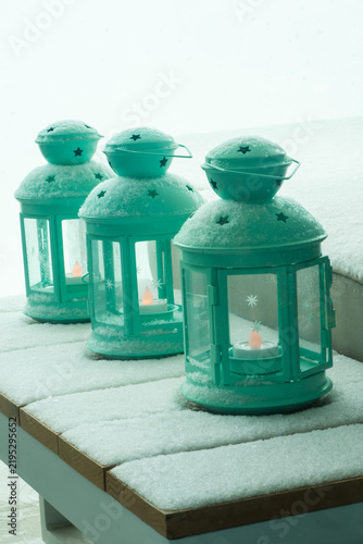 Quadro su tela Three teal metal lanterns with star-shaped cutouts and lit tealight candles stand in a row on a snow-covered wooden table, their domed tops dusted with fresh snow