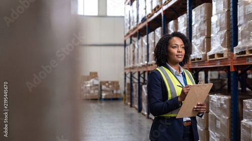 Confident African American female warehouse manager in a safety vest holding a clipboard and conducting inventory inspection in a large distribution center.