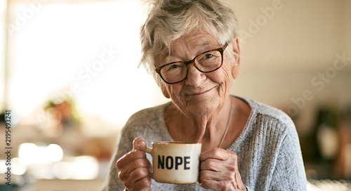 A smiling elderly woman, dressed in white, holds a humorous mug with the word 