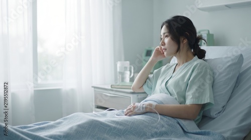 A thoughtful young asian woman patient resting in a hospital bed, looking out the window with a contemplative expression