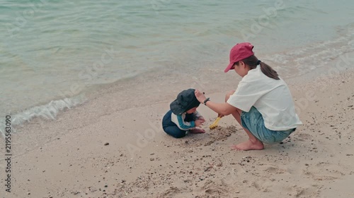 Happy mother and adorable baby boy sitting on sandy beach together during summer vacation, slow motion