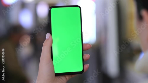 Hand holds smartphone with green screen. Background blurred with people. Three keyframes show same scene. Hand steady and centered. Urban environment visible behind