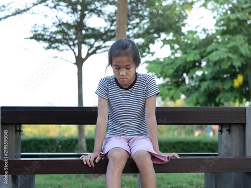 Sad and lonely little Asian girl sitting alone on park bench with depressed expression looking down
