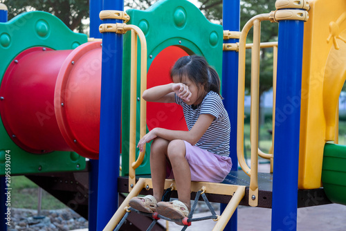 Sad little Asian girl crying and wiping tears sitting on colorful outdoor playground equipment